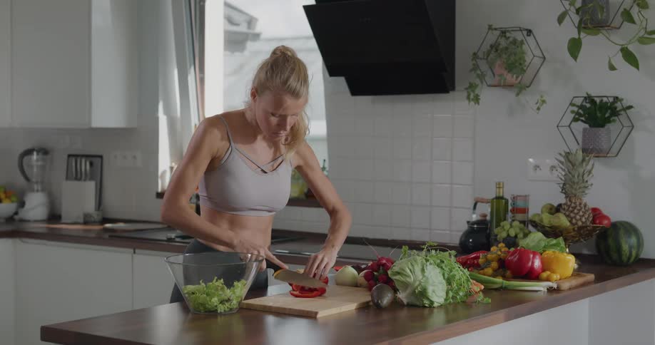 Woman wearing sports bra slices vegetables on chopping board in kitchen