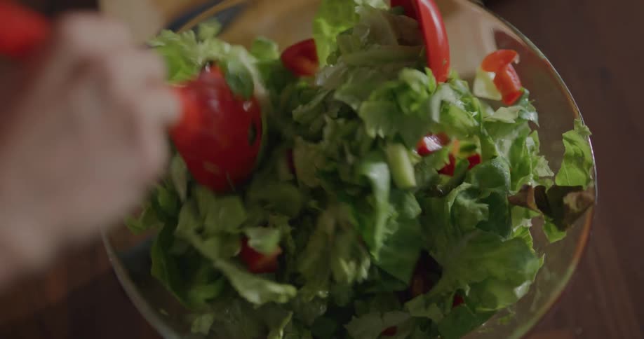 Person tosses salad in glass bowl on wooden table near chopping board and knife