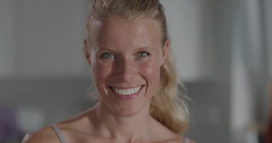 Blonde woman with a ponytail and sport bra stirs salad and stares at the camera smilingly in the kitchen