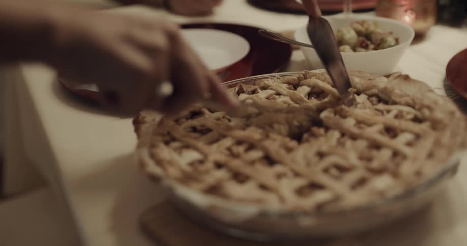 Older woman stands to slice pie on dinner table near window as people wait