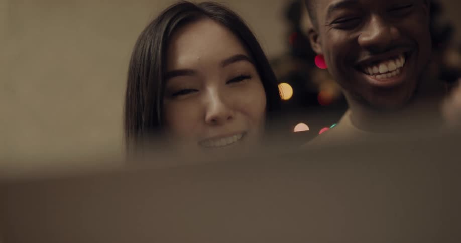 Couple smile and laugh as they talk via laptop video call near christmas tree