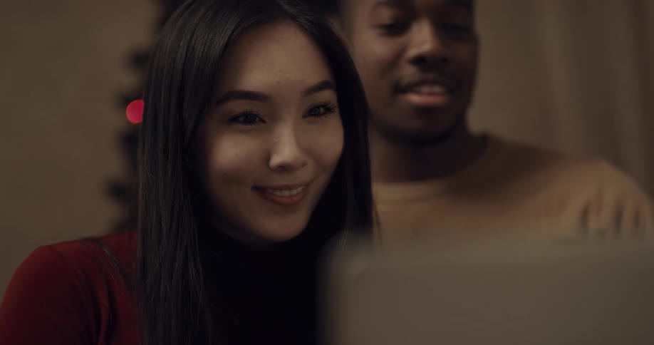 Couple smiles as they talk via laptop near christmas tree