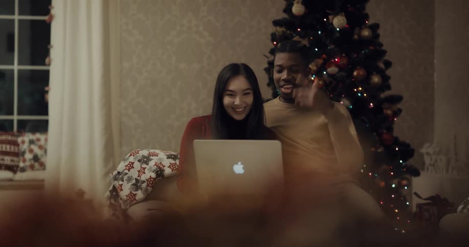 Couple sits together on chair in front of christmas tree talking via laptop