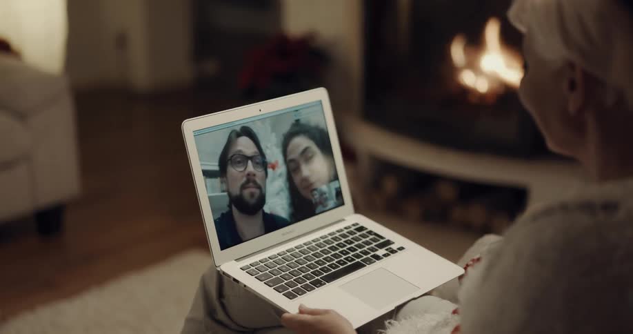 Woman with short grey hair sitting in an armchair next to the fireplace makes a video call on her laptop
