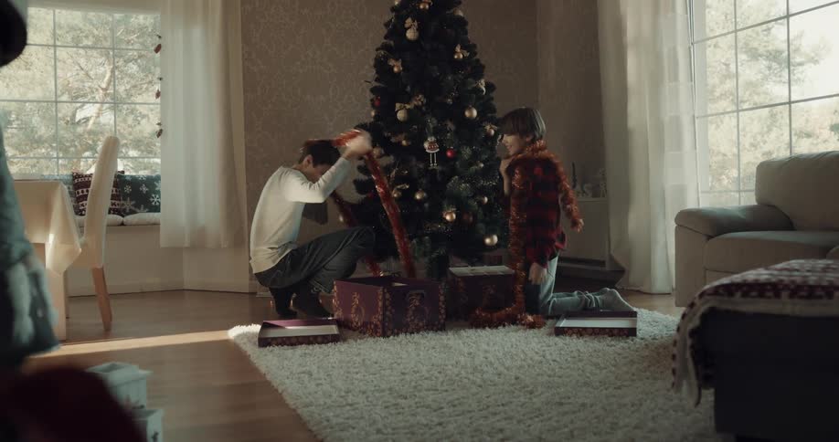 Two brothers open a box and place scarfs around their necks and ornaments on a christmas tree
