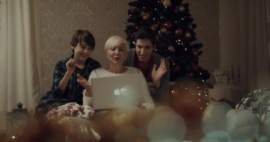 A woman and two boys in a room waving at a laptop with a christmas tree in the background