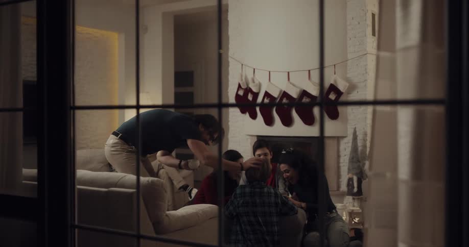 Man stands to ruffle hair of boy sitting on ground with others in living room near fireplace with christmas stockings