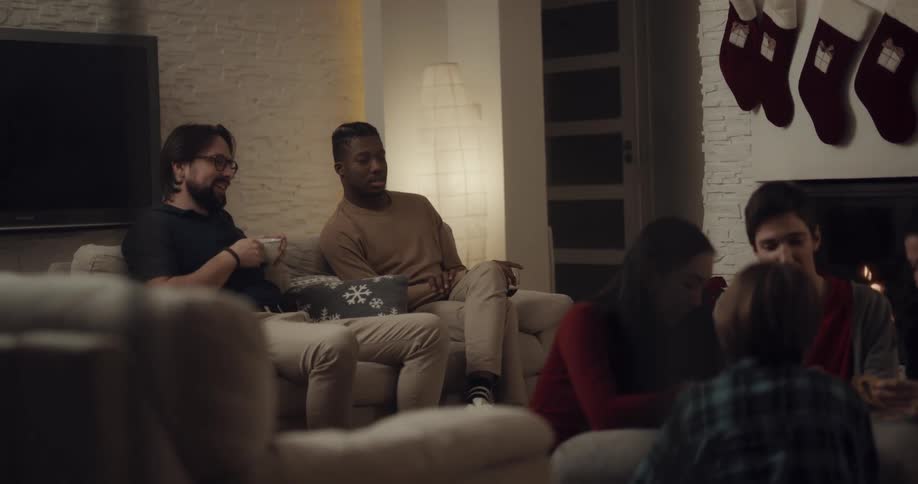Two men sitting on couch talk while watching children play game at coffee table near fireplace