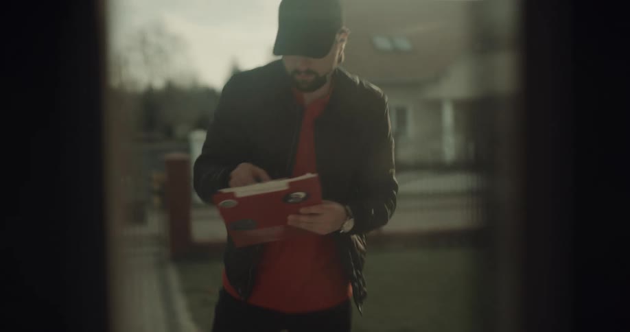 A bearded man wearing a baseball hat delivers boxes to the front door of a house and marks off on a clipboard being carried