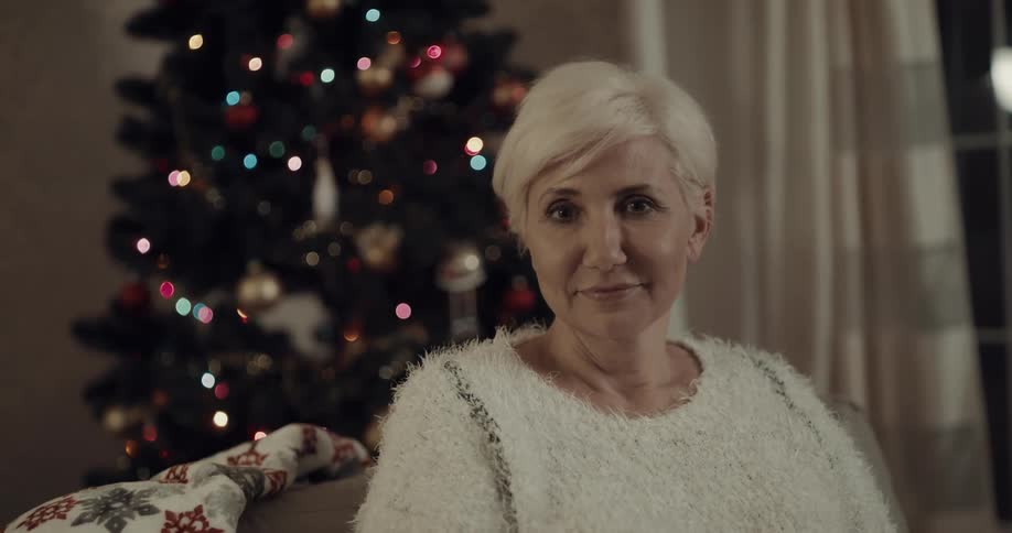 Older blonde woman sits on couch next to christmas tree near window showing night
