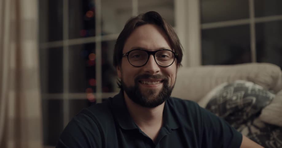 Man with glasses beard and a black polo shirt looks at the camera smilingly in the living room at home