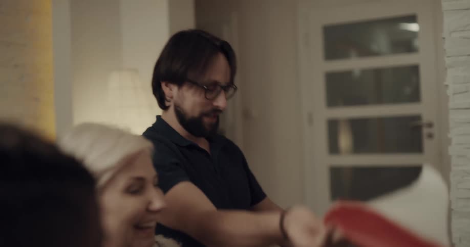 Older woman and others laugh as man opens gift sitting near fireplace