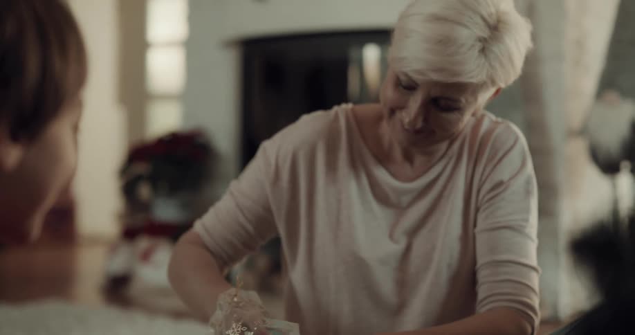 Boy hands older woman gift while they sit on floor in living room near christmas tree