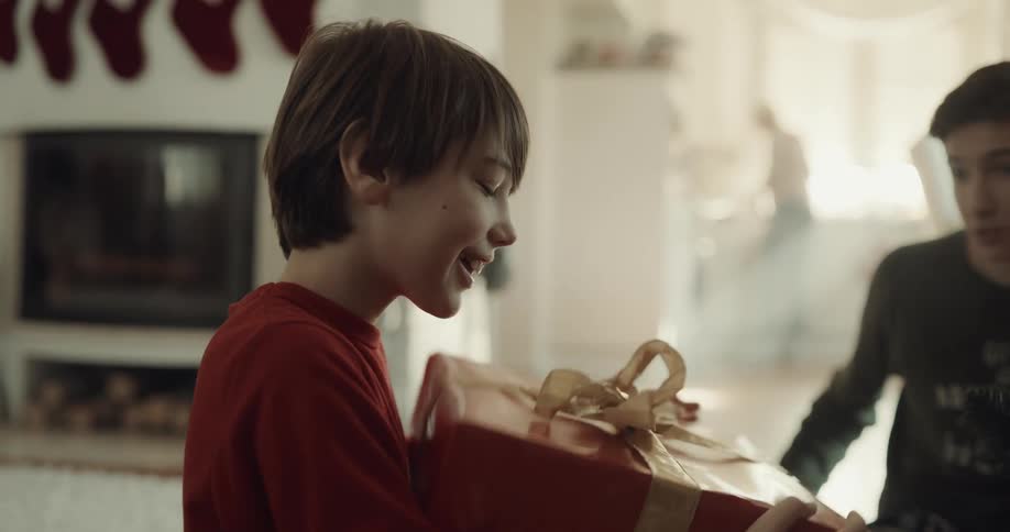 A boy sitting in front of a christmas tree retrieves a large present from beneath the tree and begins to unwrap it