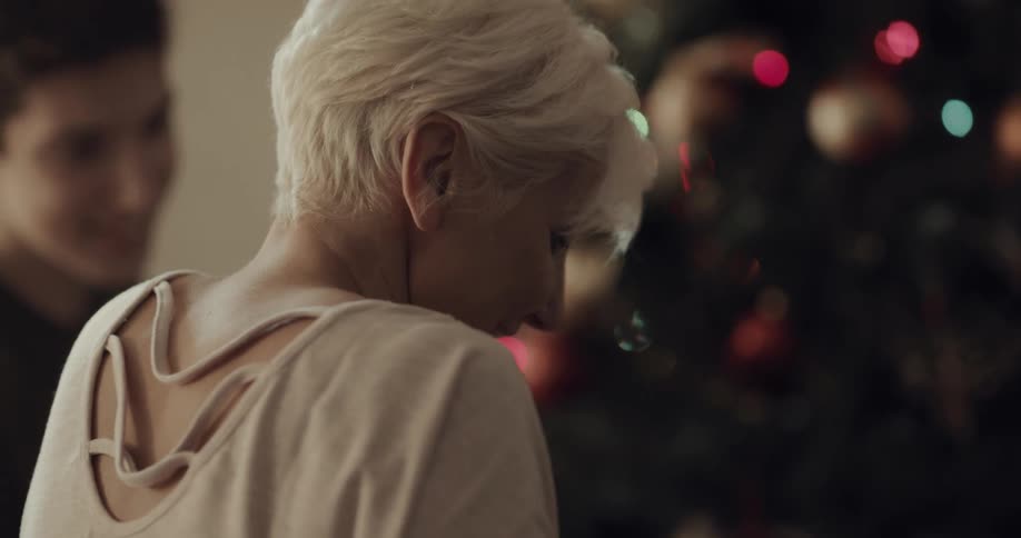 A woman opens a gift box next to her teenage grandson and a christmas tree