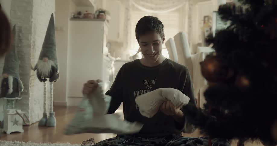 Brothers open presents sitting on floor near christmas tree as older woman works in kitchen