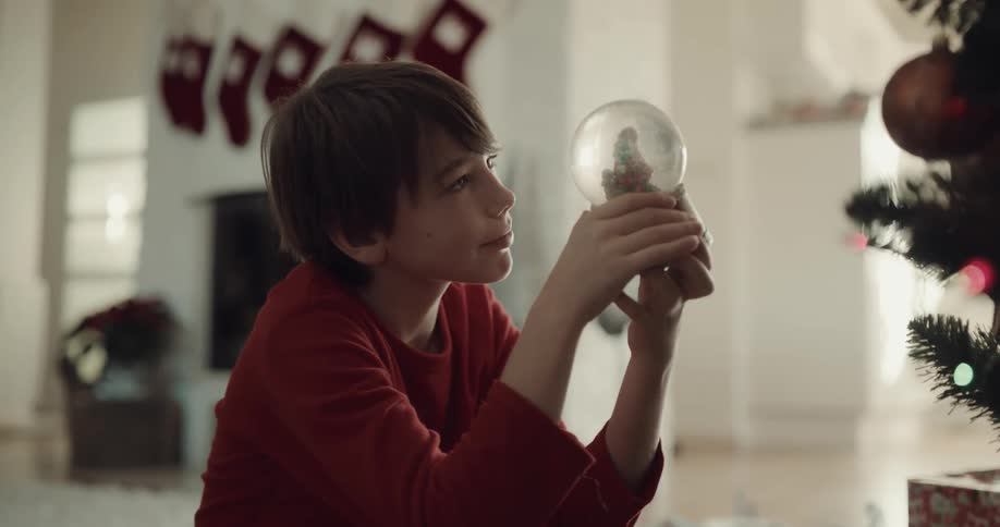 A boy sits by a christmas tree at home and shakes a snow globe
