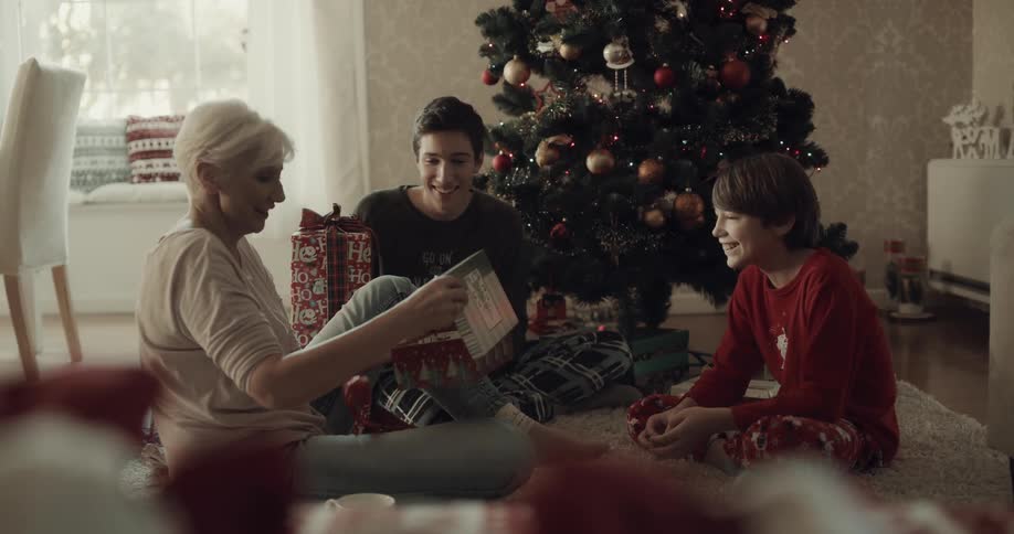 Woman with short grey hair sitting on the ground next to the christmas tree and unwraps a present and kiss her grandsons