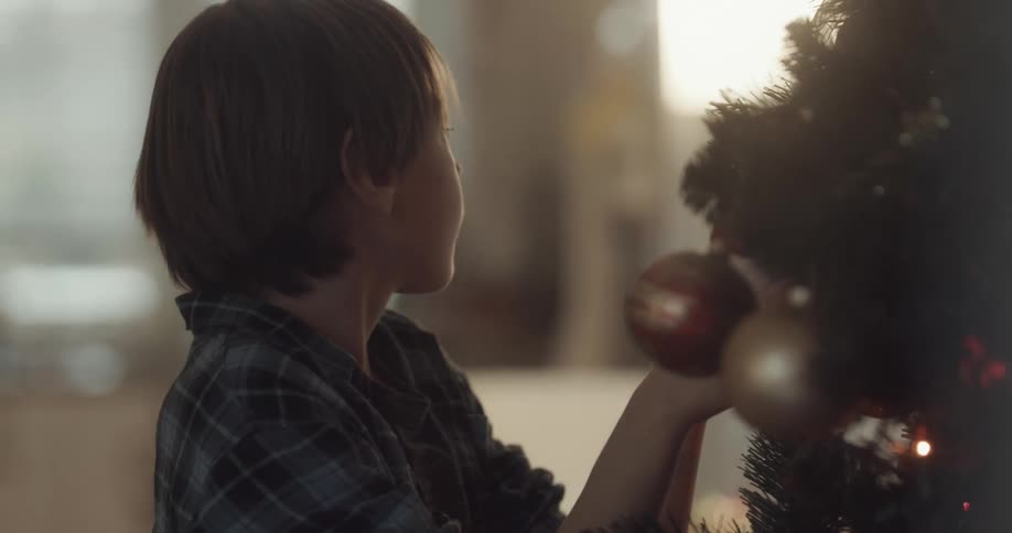 Boy decorates christmas tree with ornaments as older woman moves around in background