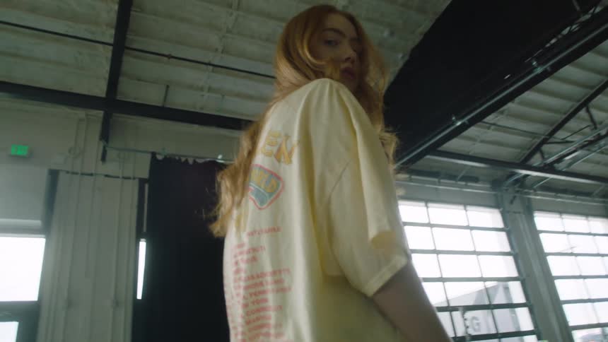 Model with red hair wearing jeans and a t shirt walks toward photography equipment and lights looking over her shoulder at camera inside a metal warehouse building with rafters overhead and windows and an exit sign in the background