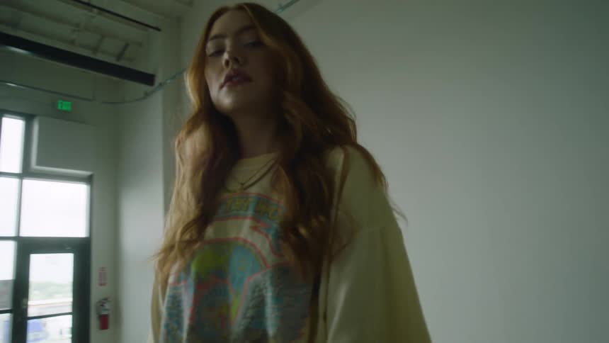 Model with red hair stands in front of a white wall wearing a t shirt jeans and a necklace holdingjacket beside a door windows a fire extinguisher and an exit sign