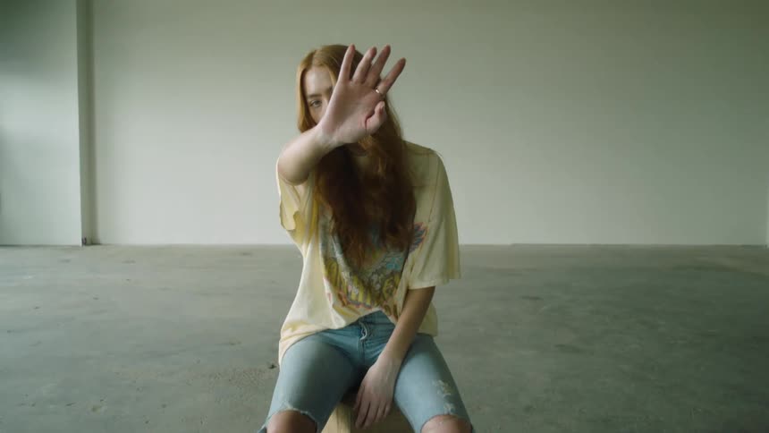 A teenage girl sits in an empty warehouse and holds her hand in front of her face and sways side to side
