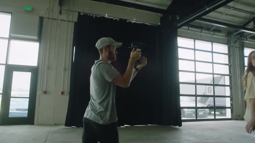 Photographer takes pictures or records a model walking and posing in front of high windows and black curtains with cars and trucks outside in a warehouse building with a metal roof and rafters