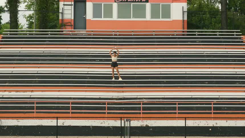 A woman stands outside midday on some bleachers stretching and prepping for athletic training