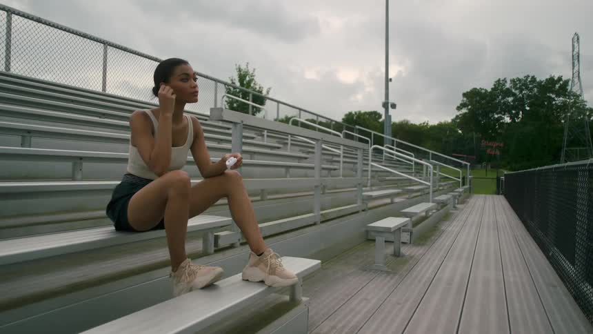 A young woman puts ear buds in her ears while sitting in the otherwise empty bleachers of an outdoors athletics field on a cloudy day