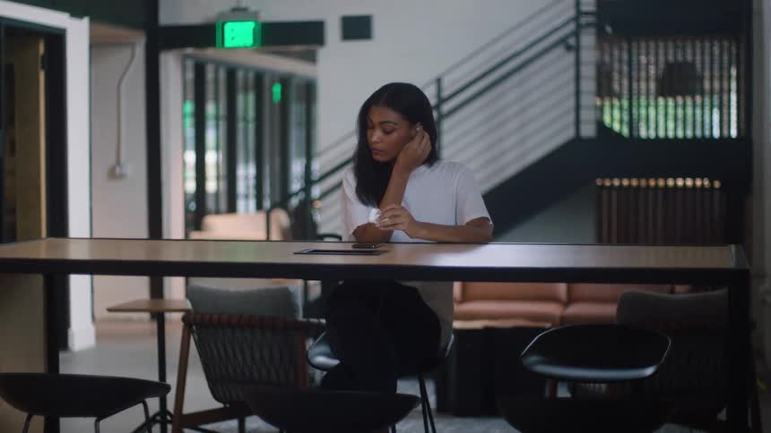 A young black woman sits at a table in a building and puts airpods in her ears