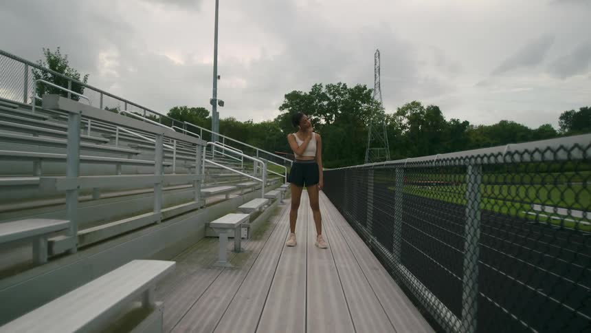 A woman stretches her neck while standing in workout gear next to bleachers at a track stadium