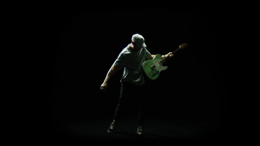 A man poses with his guitar and spins around in a dark studio