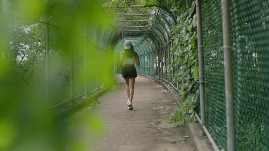 Woman with long hair wearing running shoes shorts a tank top and a hat runs down a fenced in concrete path with ivy growing on it and trees in the background