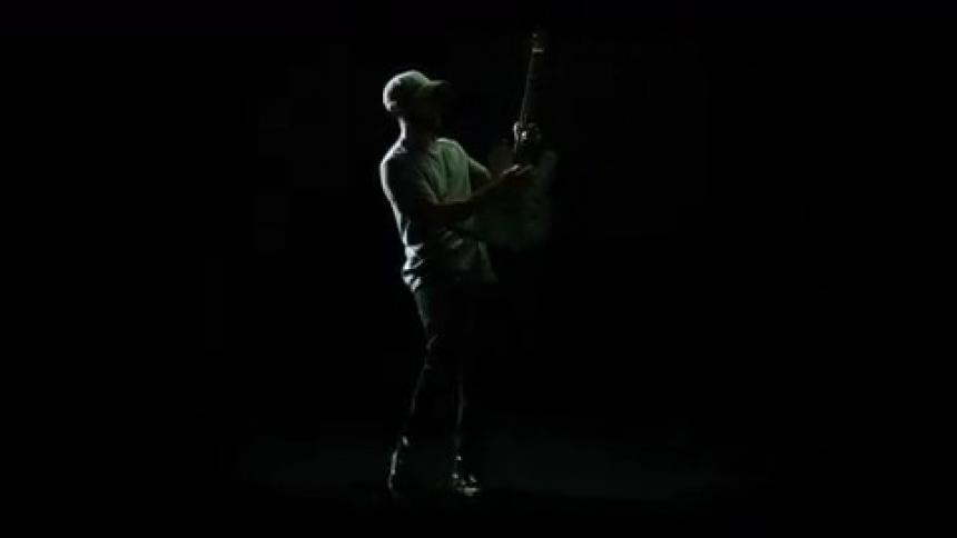 A man stands in a pitch black room with flashing lights playing a guitar in a jerking motion