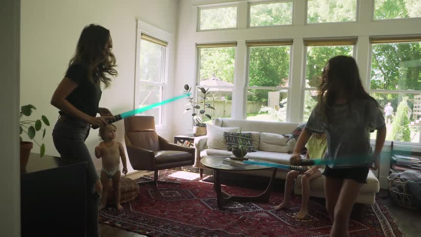 Mother and daughter play sword fight with light sabers in living room while two other children watch one child one toddler
