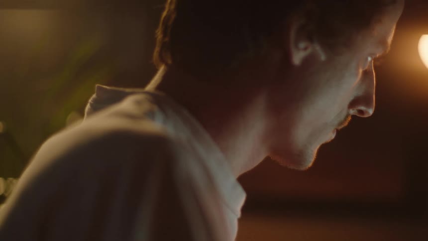 A man wearing a white and watch sits looking at something in thought as he twitches his fingers in a room lit by a light fixture
