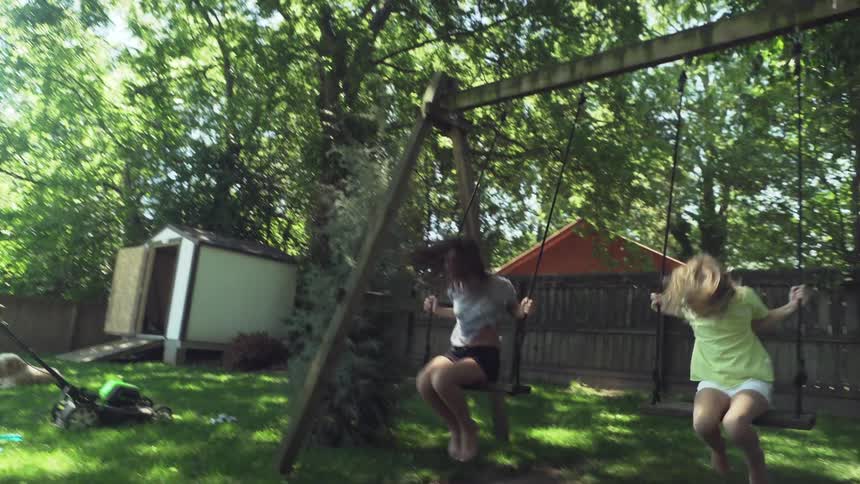 Two girls swing higher and higher on swing set in backyard until one jump off