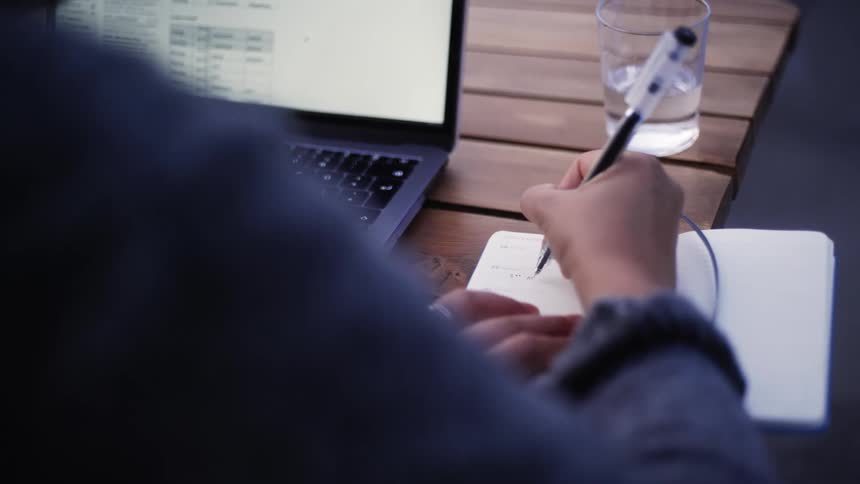 A person writes in her notebook while using a laptop