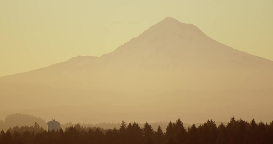 A mountain sits behind and above a forest area and hills with a small building among the trees