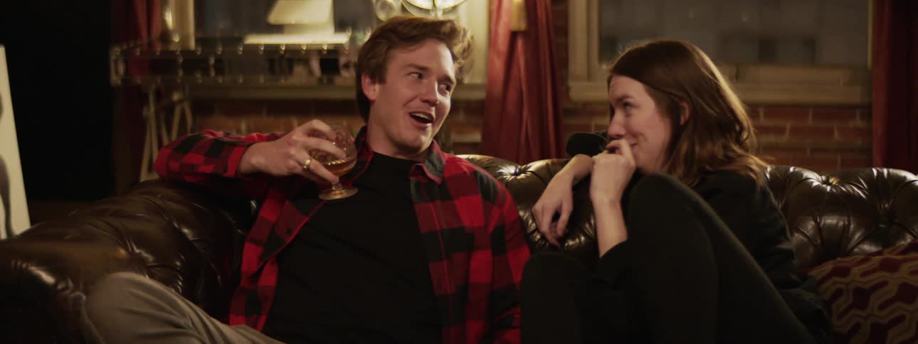 A man and woman smile and laugh as they sit on a couch while drinking alcohol