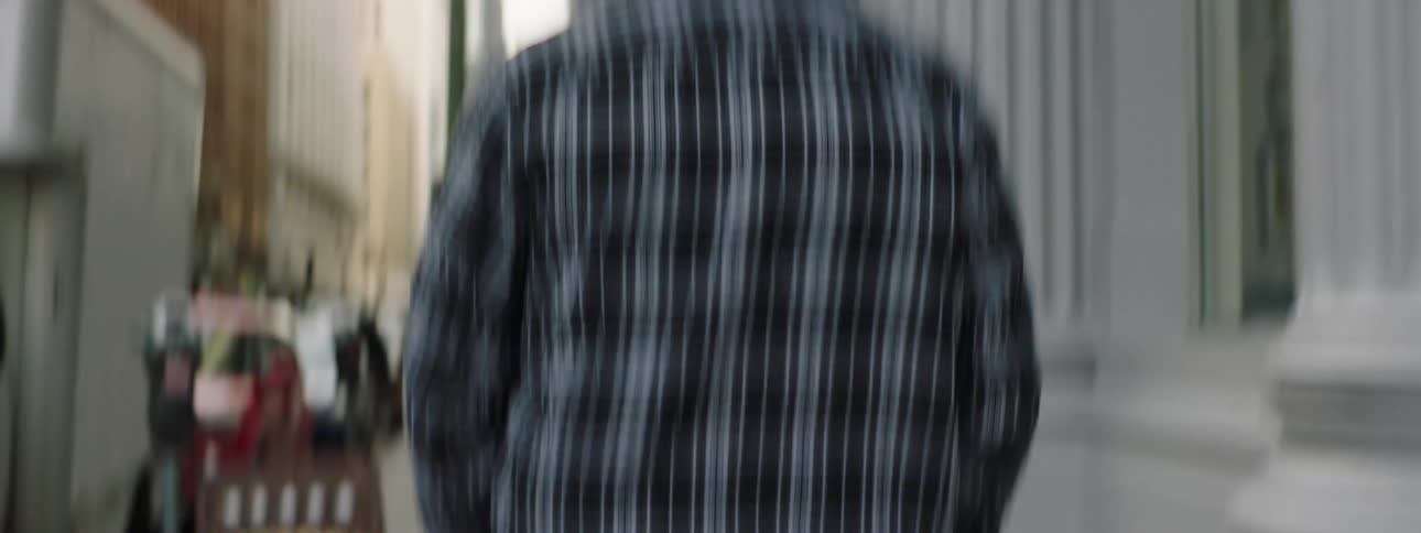 Man wearing brown shoes jeans and a plaid long sleeved shirt walks on sidewalk of city street past buildings signs a potted plant and a truck and cars in the background there is a slip of sky more buildings street lights ivy on the side of a building