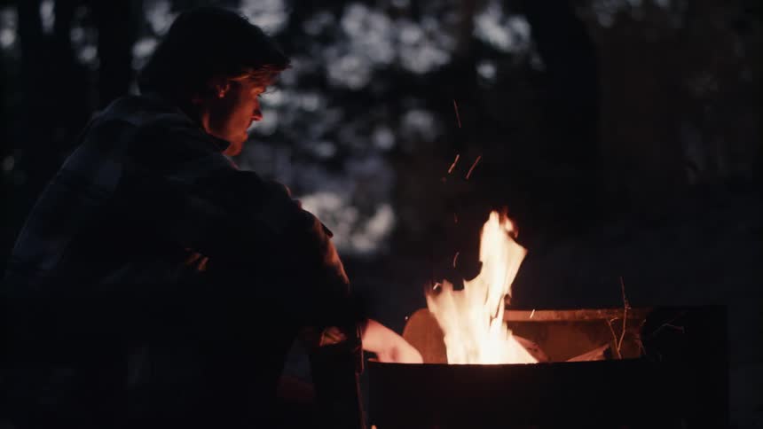 A man sitting next to a fire and adding a log to the flame with trees the sky and clouds in the background