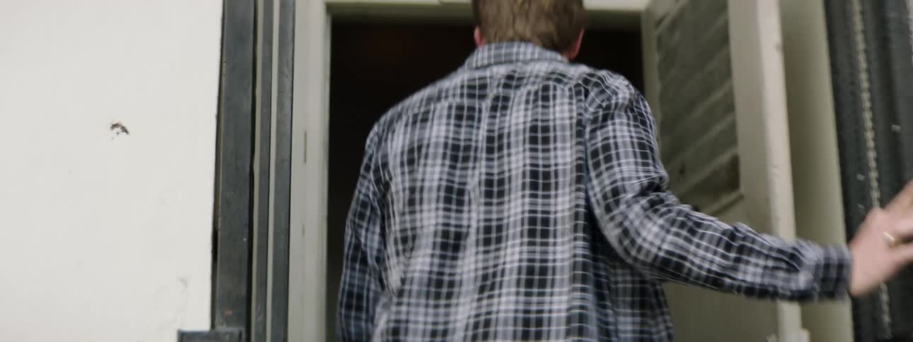 Man waring long sleeved shirt pants and a ring walks to and opens a screen door then walks through already open metal door into building
