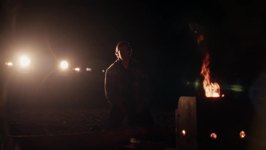 A man with a jacket kneeling down with a fire in the foreground and lights in the background