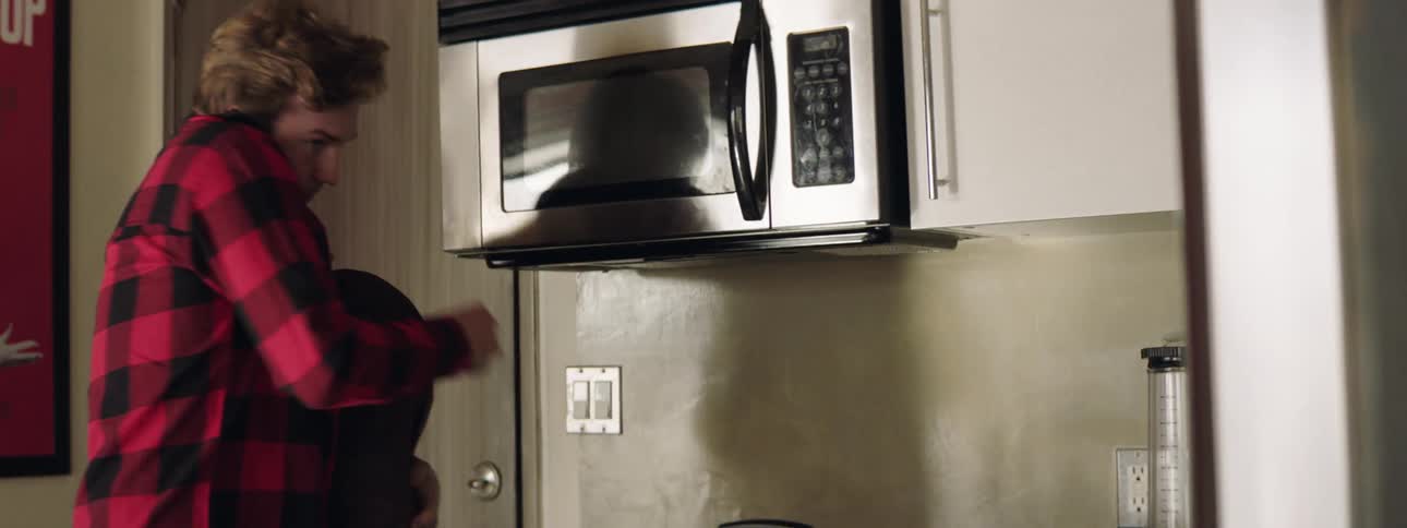 Man comes in door wearing plaid red long sleeved shirt carrying grocerieswhich he moves to put in the refrigerator shuts door there is a poster a microwave and light switches on the white walls there is a stove with a tea kettle on top