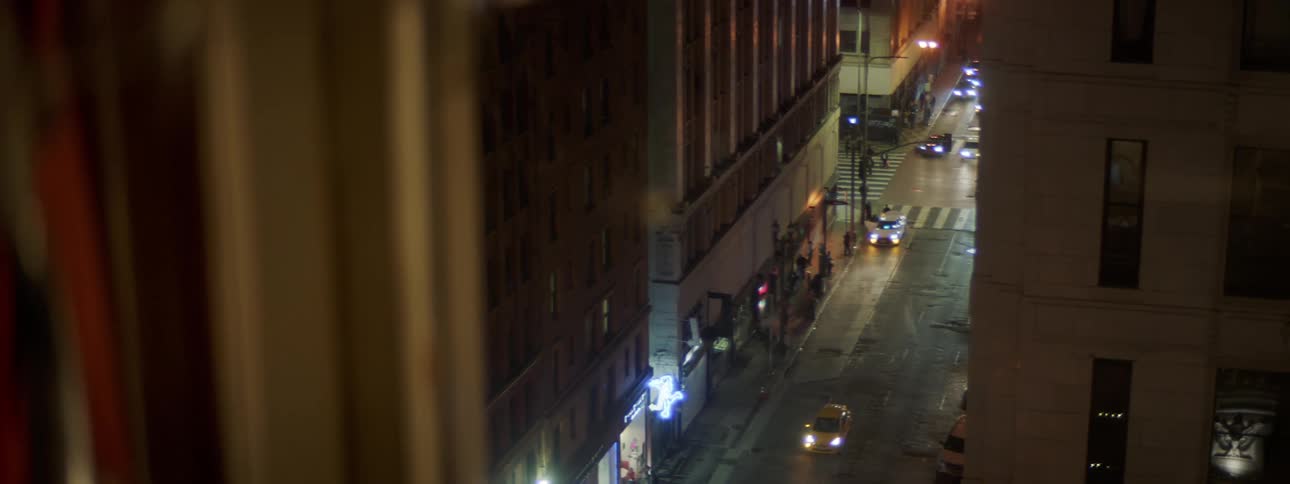 From an apartment window vehicles drive down a city street at night past high rise buildings and flashing lights