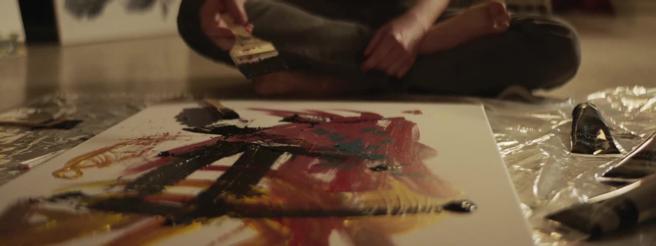 A man sits on the floor painting on a large canvas with a plastic tarp underneath