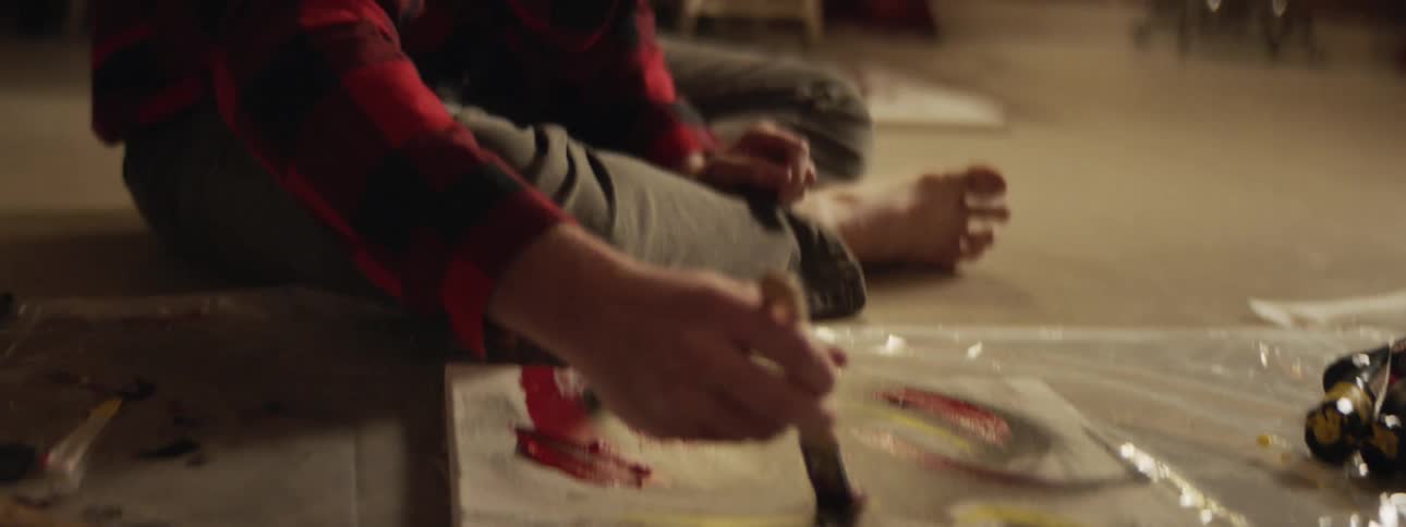 A man sits on the floor painting on a canvas with a plastic tarp underneath