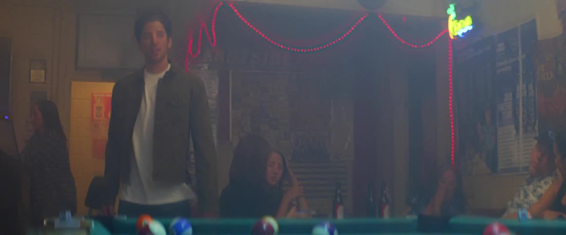 Man walks into a room and towards a pool table while a pool player hits a ball with a pool cue