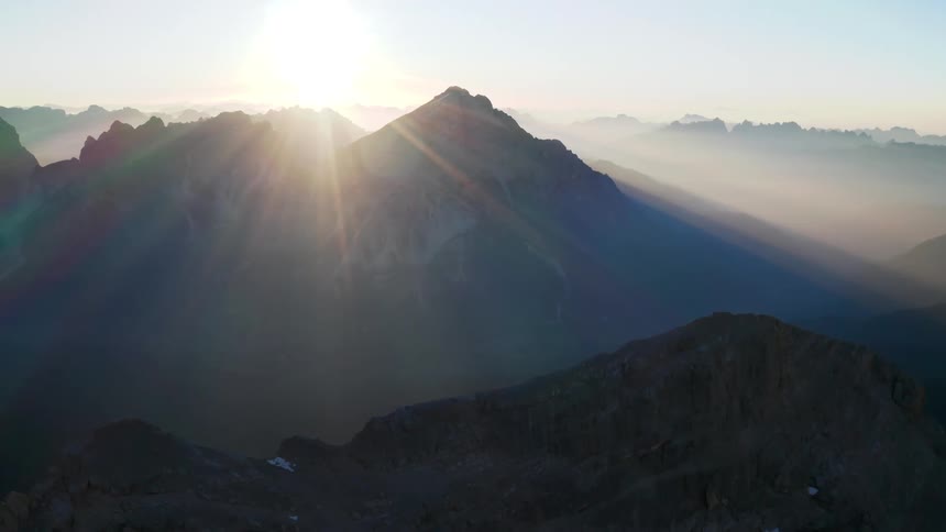 The suns rays shine brightly onto foggy mountain ranges with moderate peaks and little snow